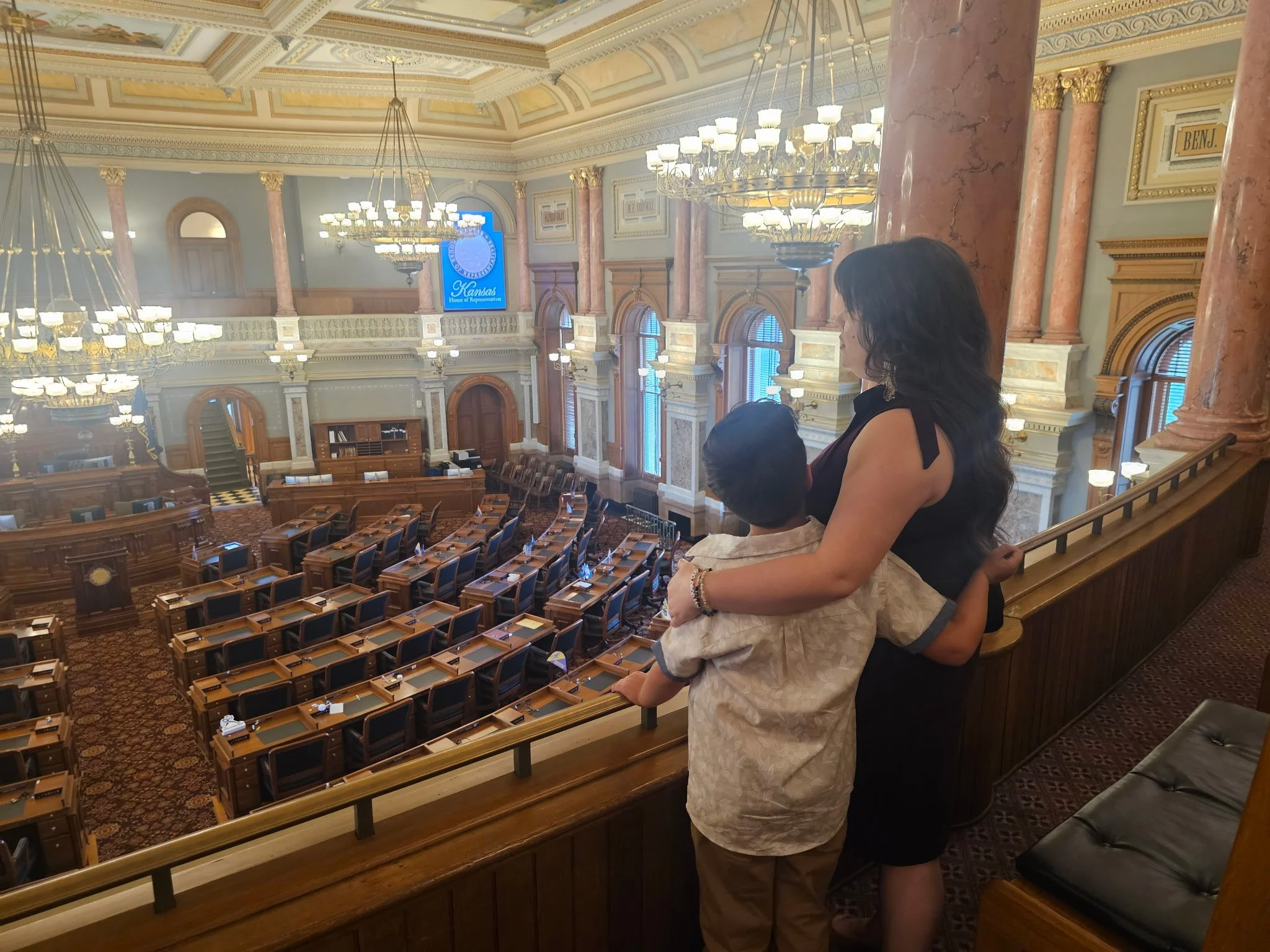 Interior of a government chamber with ornate decor, chandeliers, and a balcony. Two women are observing from the balcony, one with black hair wearing a dark top, and a young boy with dark hair wearing a light shirt.