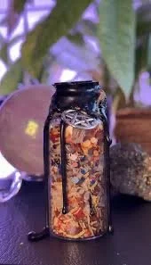 A glass jar filled with gold, copper, and silver coins, with a black lid and a black string tied around its neck, placed on a dark surface with plant leaves and a spherical object in the background.