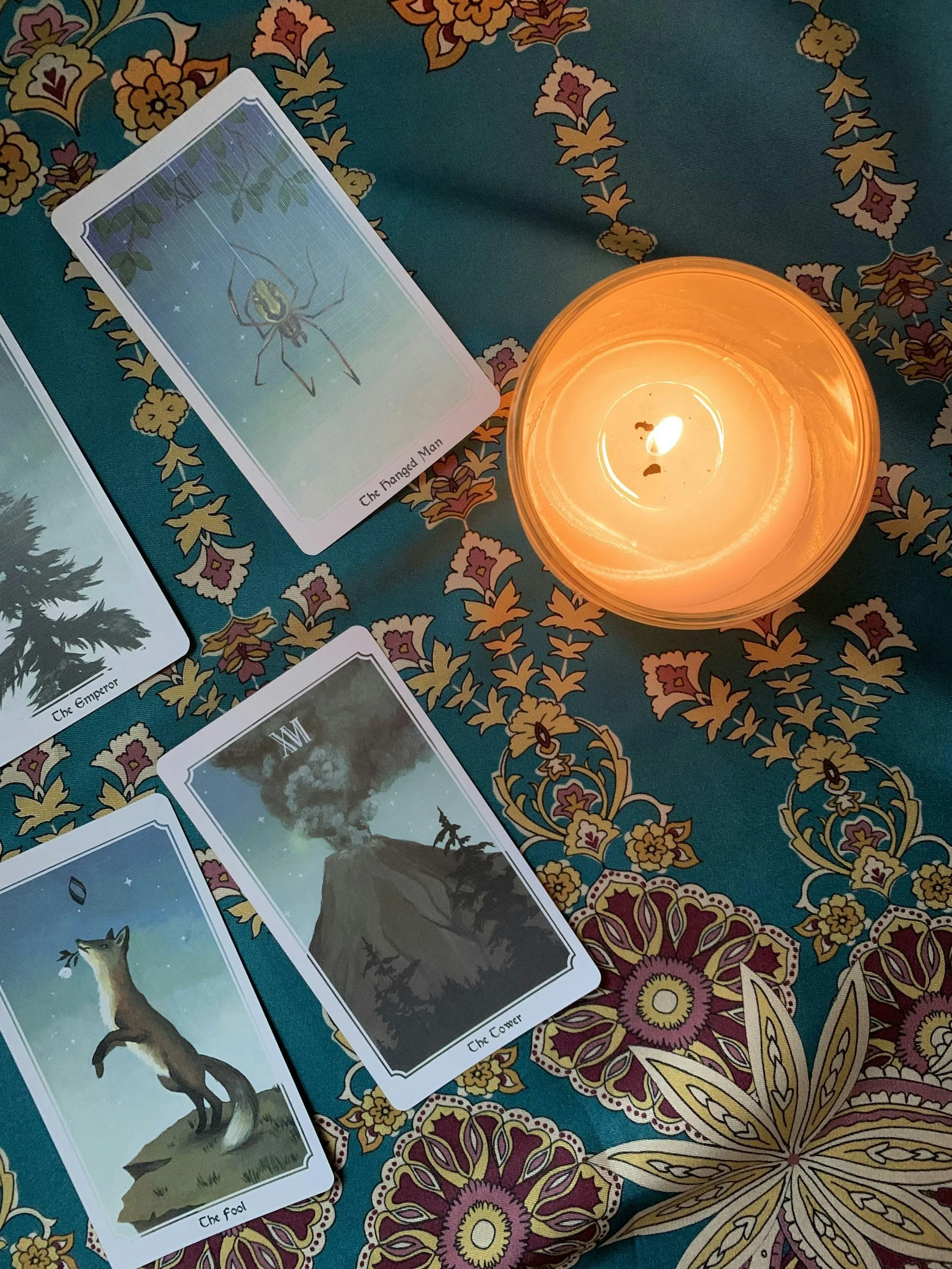 Tarot cards laid out on a colorful, floral-patterned tablecloth, with a lit orange candle beside them. The visible cards include 'The Fool,' 'The Empress,' and 'The Tower.'