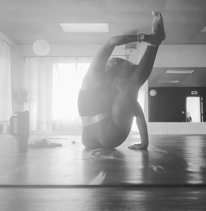 Person doing a yoga pose indoors, balancing on one hand with legs bent over their head, in a room with large windows and curtains, black and white photograph.