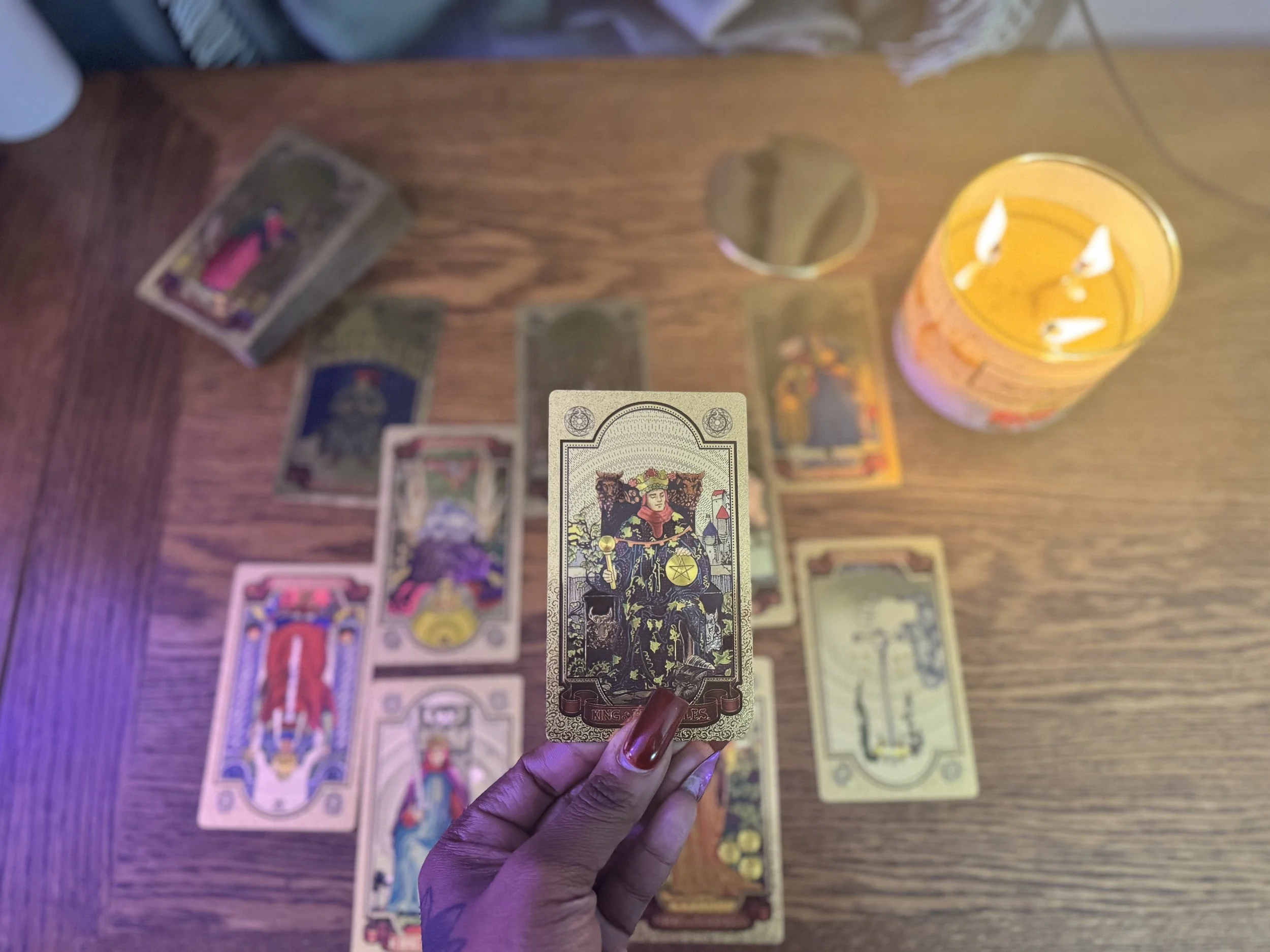 Close-up of a hand holding a tarot card with a woman in a black dress and two lions on either side, surrounded by other tarot cards laid on a wooden surface, along with a candle and various tarot cards in the background.