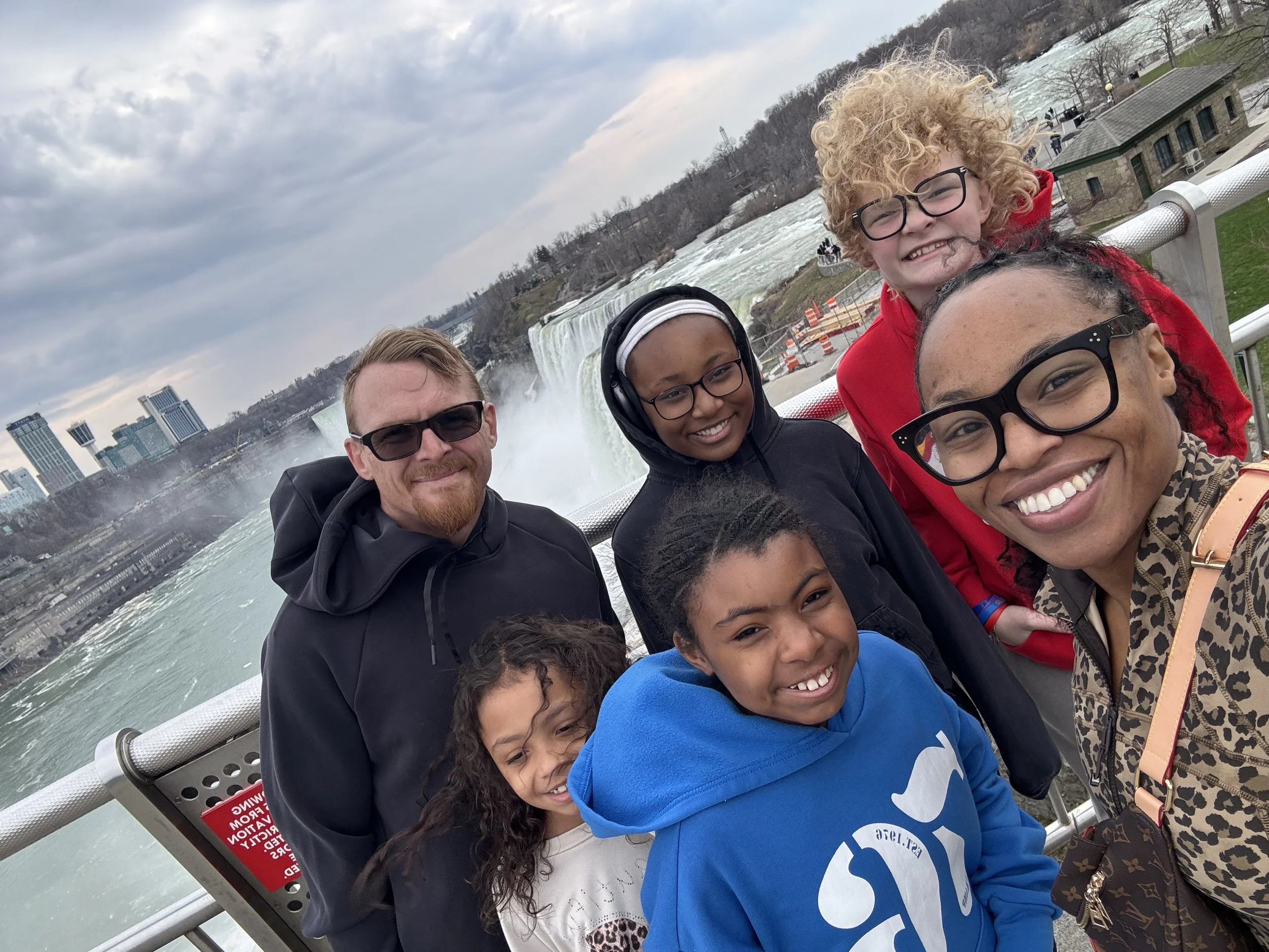 A group of smiling people taking a selfie on a boat. In the background, water and waterfalls are visible, with buildings on the shoreline and cloudy sky above.