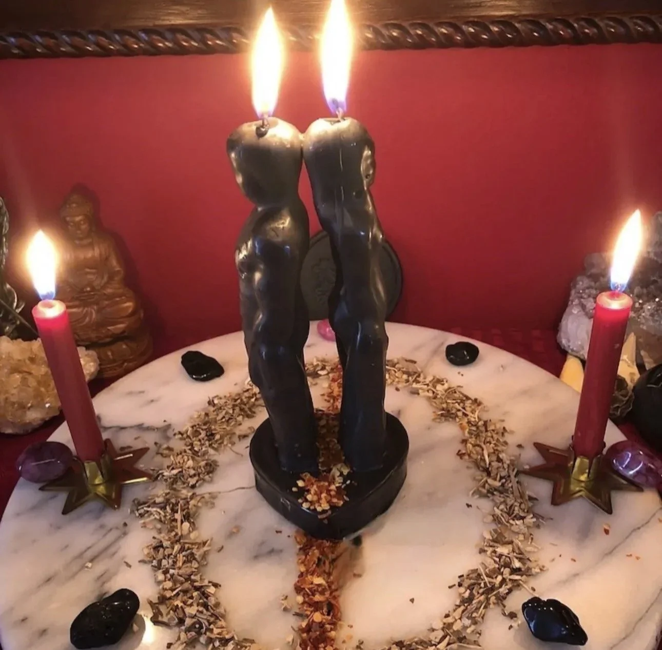 A pair of black, modern, sculptural candles shaped like two elongated, intertwined figures with flames on top. The candles are on a white marble surface decorated with a circular arrangement of dried herbs and flowers. Around the candles are small polished stones, and on each side, there are lit red candles in star-shaped holders. In the background, there are small figurines and crystals against a red wall.
