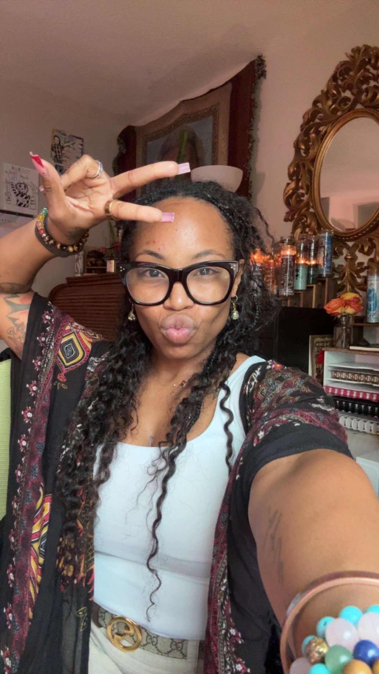 A woman with curly hair, glasses, and jewelry taking a selfie in a room decorated with various items, including a mirror, candles, and framed pictures.