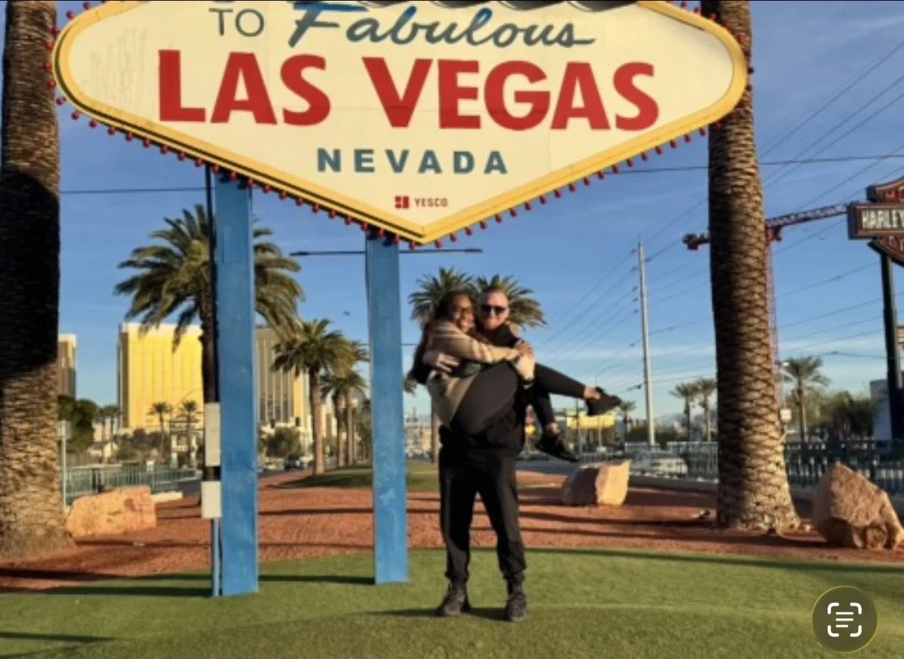 A man holding a woman in his arms under the 'Welcome to Fabulous Las Vegas Nevada' sign with palm trees and city buildings in the background.