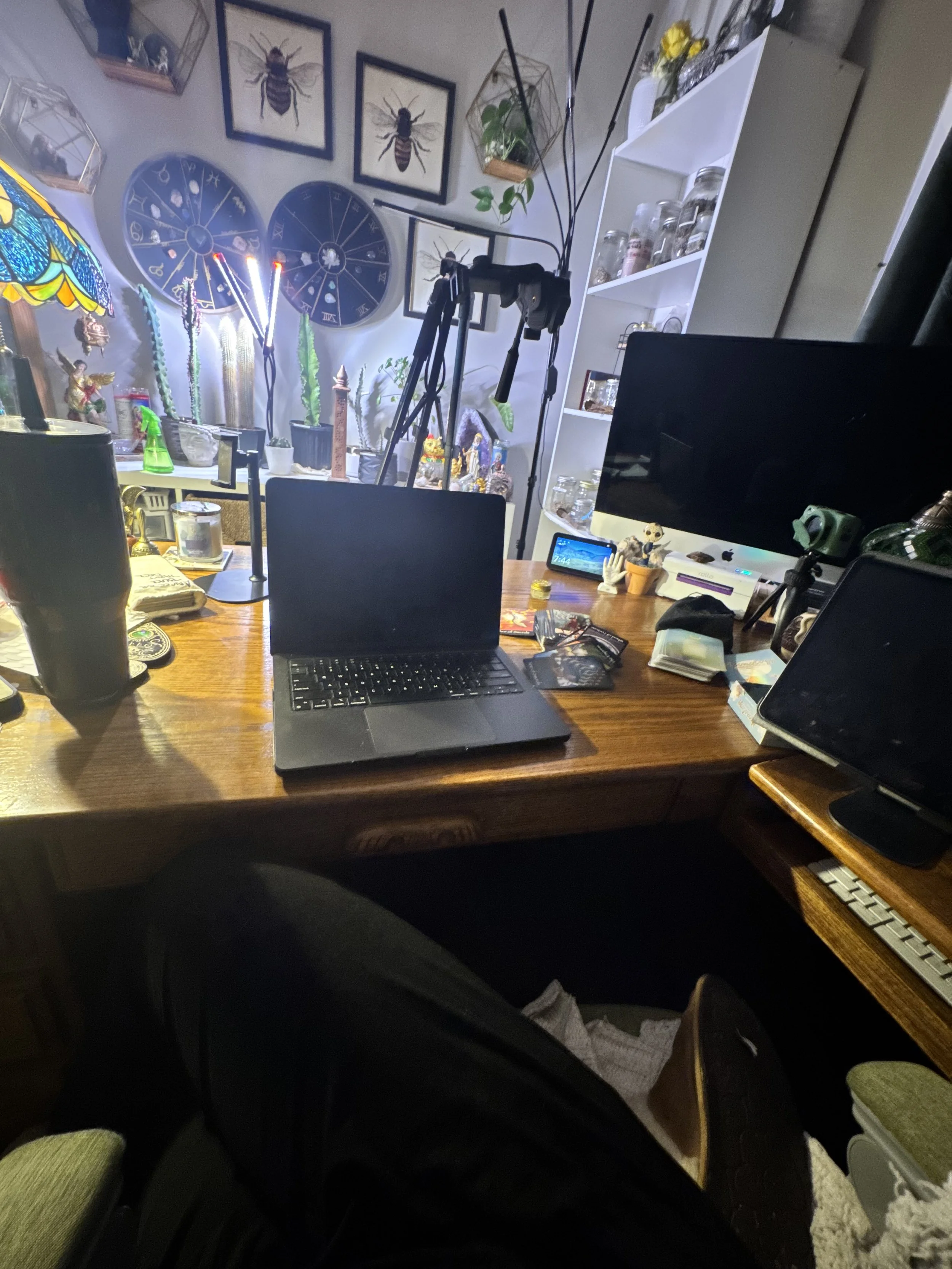 A cluttered home office or living space with a wooden desk in the foreground. The desk has a laptop, a tablet, and various papers and objects. Behind the desk, there are white shelves filled with jars, plants, and decor. The wall above features frame