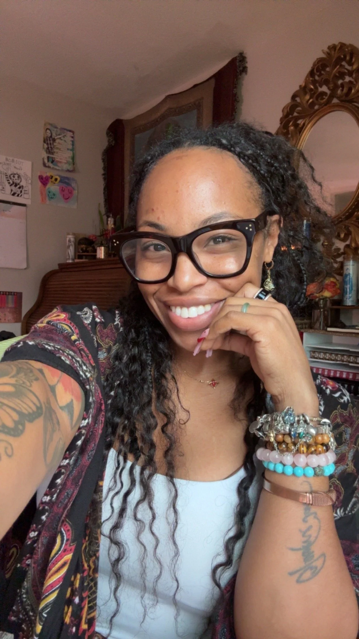A woman with long curly hair, wearing glasses, a white top, and colorful bracelets, taking a selfie in a room with a mirror and wall art.