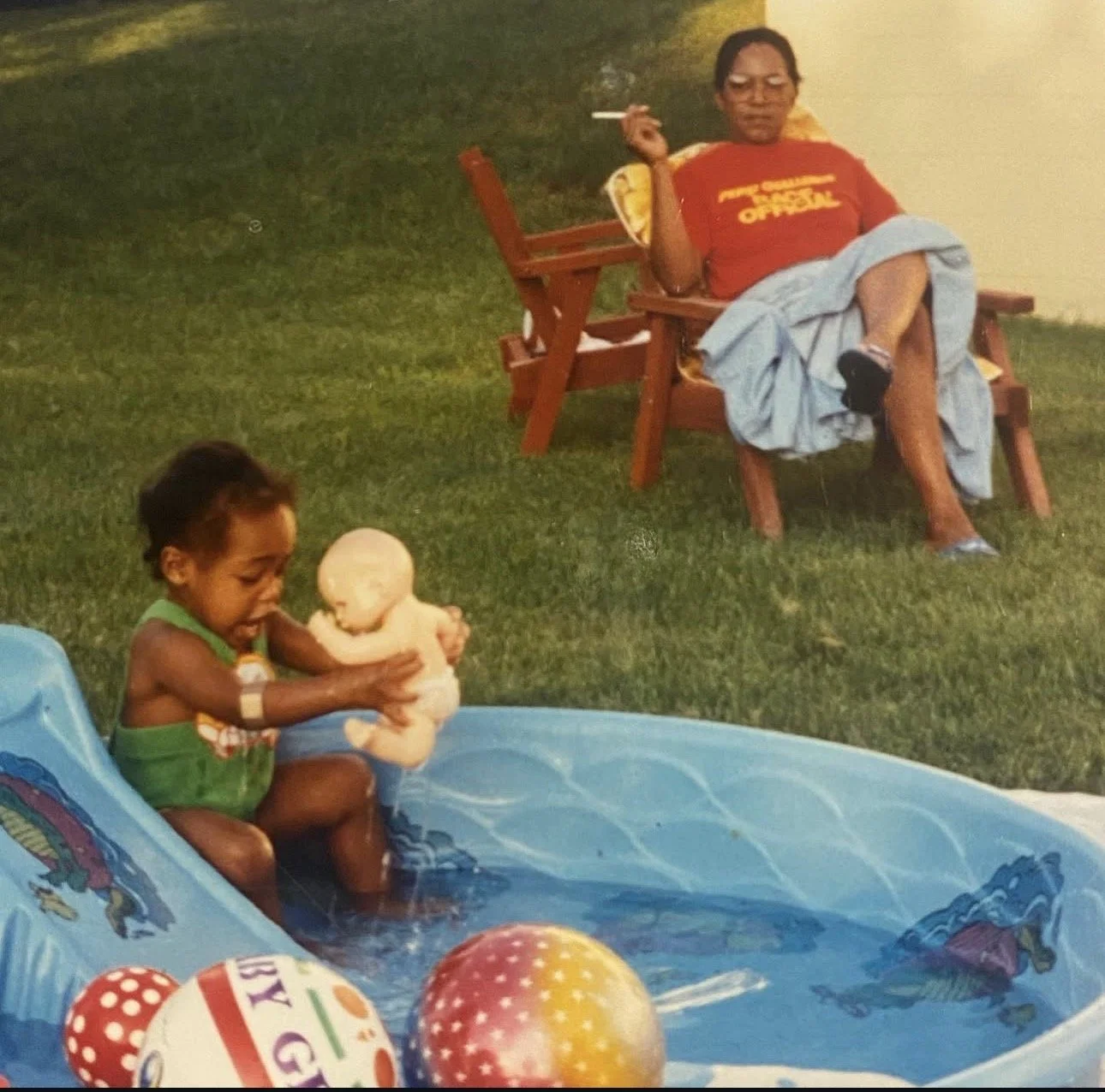 A young child playing in a small inflatable pool with a doll, while an older woman relaxes on a bench nearby with a cigarette in her hand.