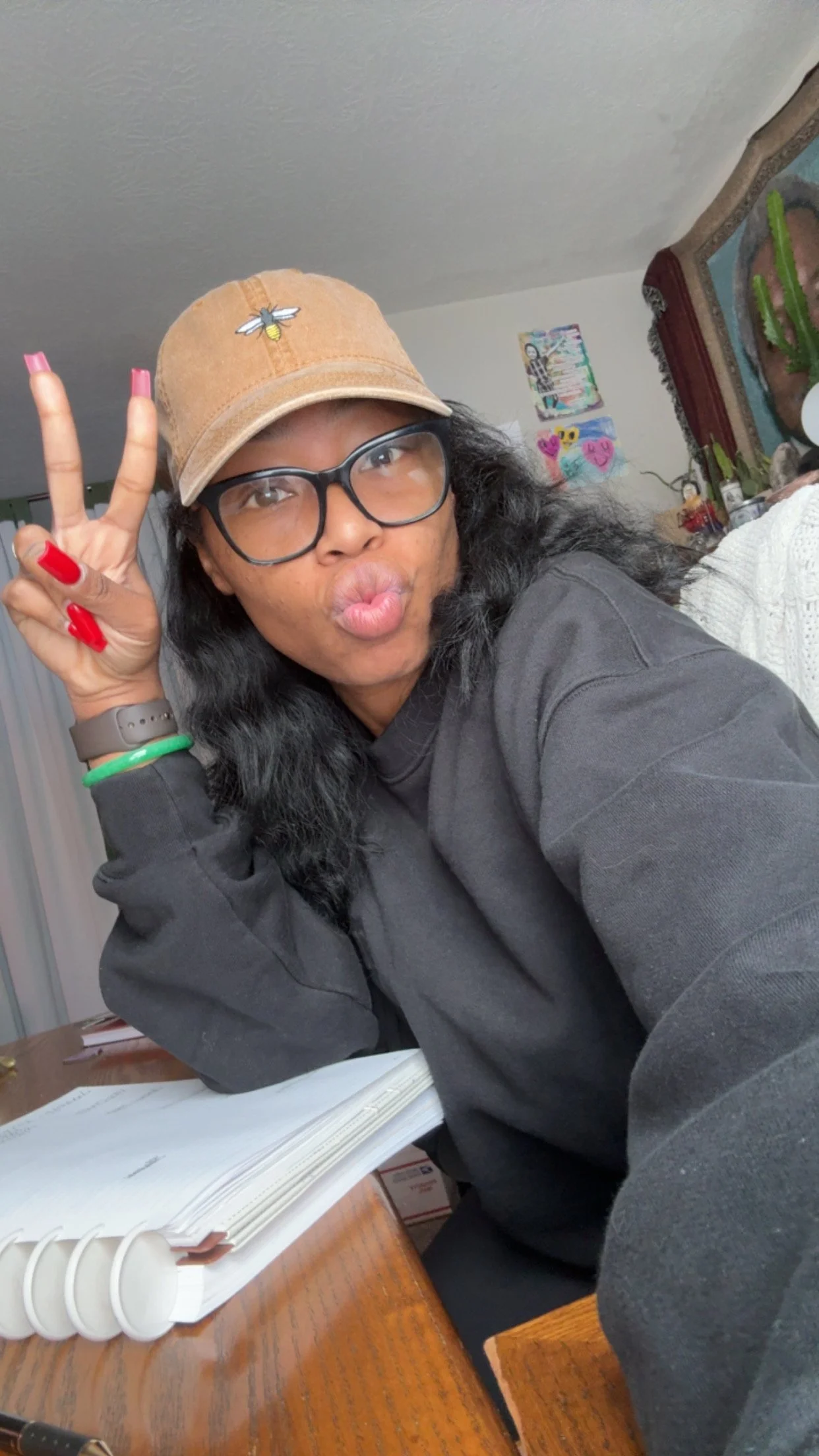 A woman with glasses and a beige cap making a kissy face and peace sign in a room with colorful art on the wall behind her.