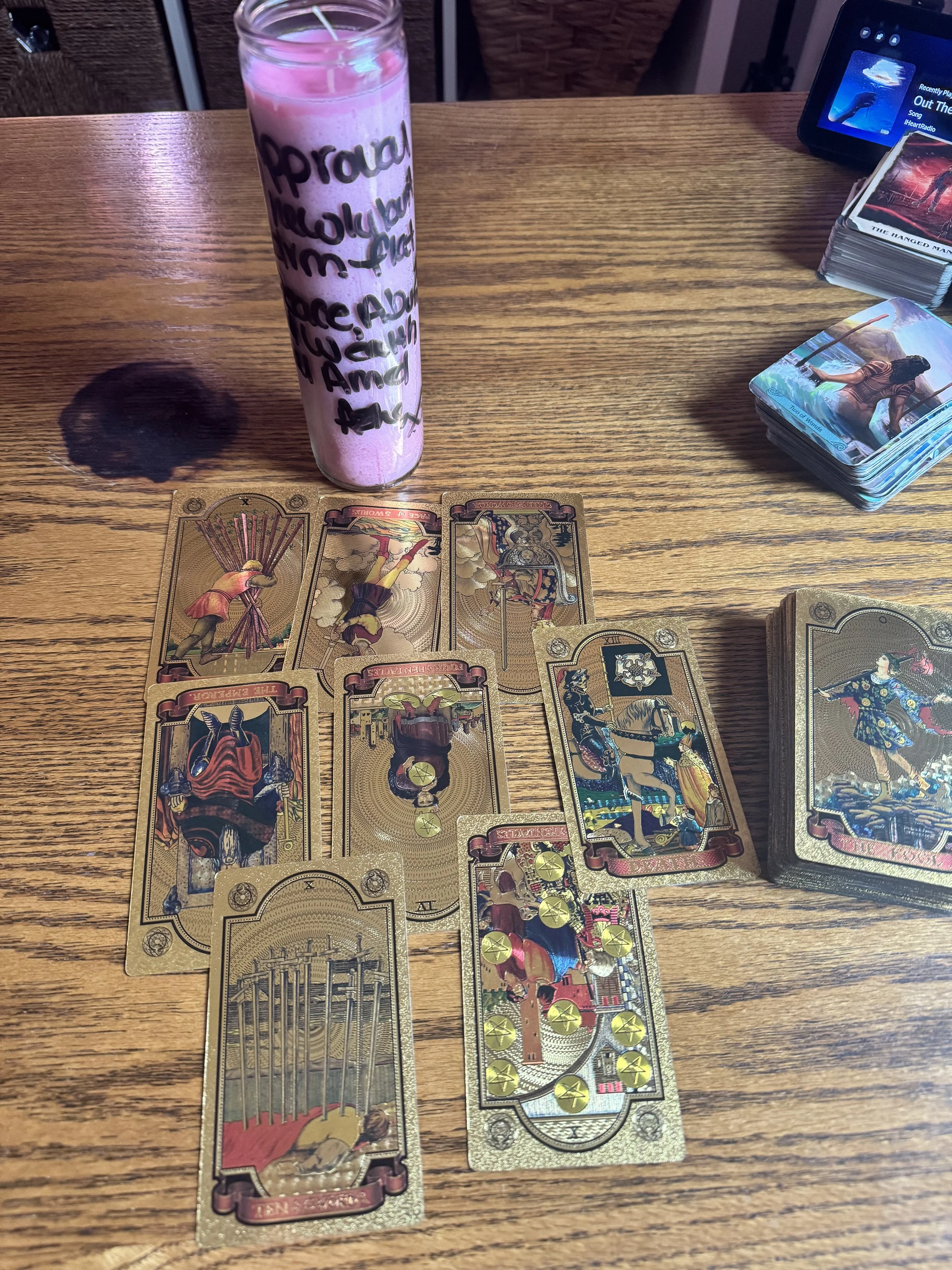 A wooden table with tarot cards spread out, a glass jar with pink liquid and writing on it, a tarot card box, a digital device, and a deck of cards with an illustration of a woman in a dress.