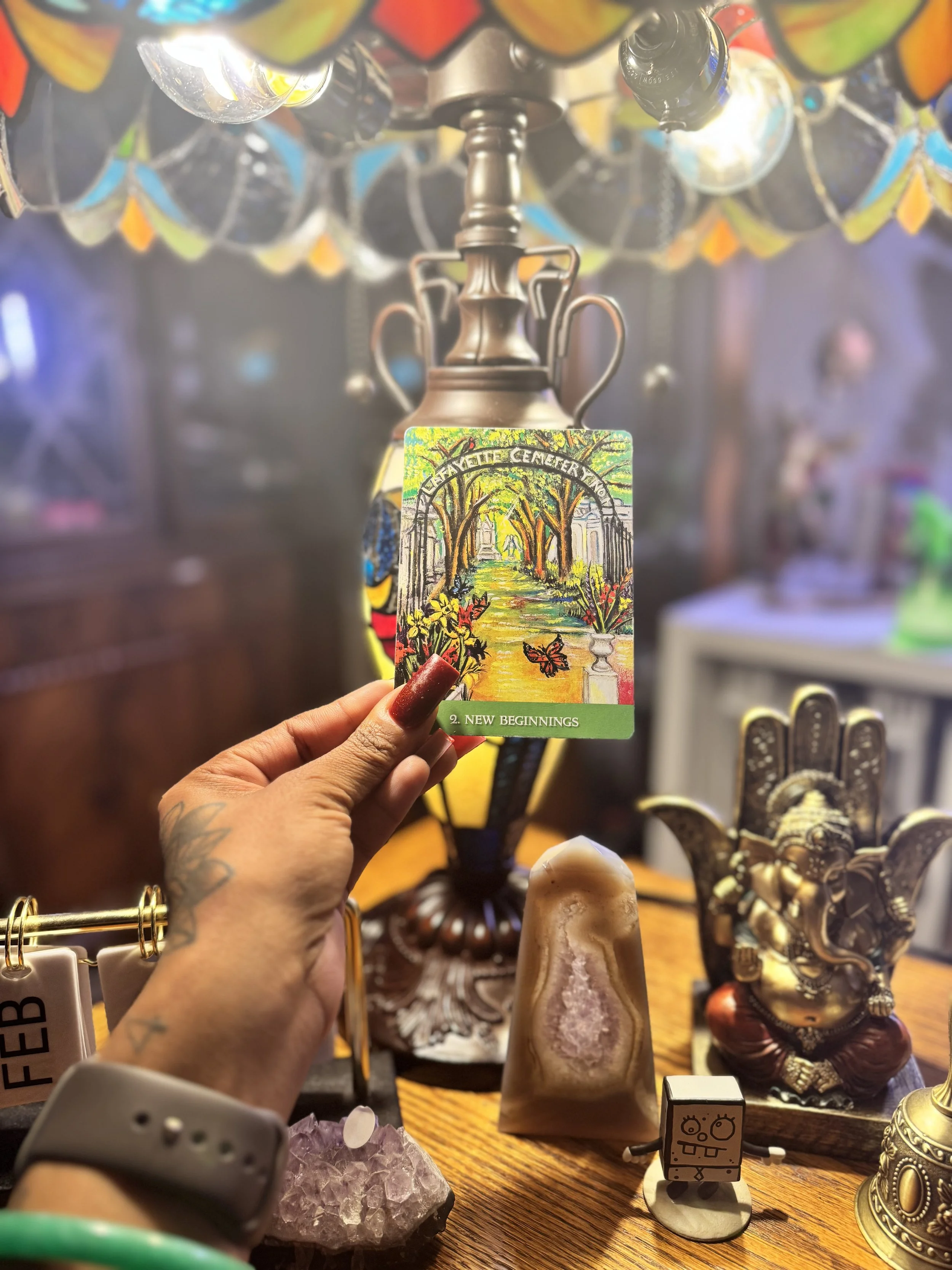A person with red painted nails holding a colorful card that says '2. New Beginnings' in front of decorative items, including a metallic figurine, a small cartoon-like robot figure, and other assorted figurines on a wooden surface.