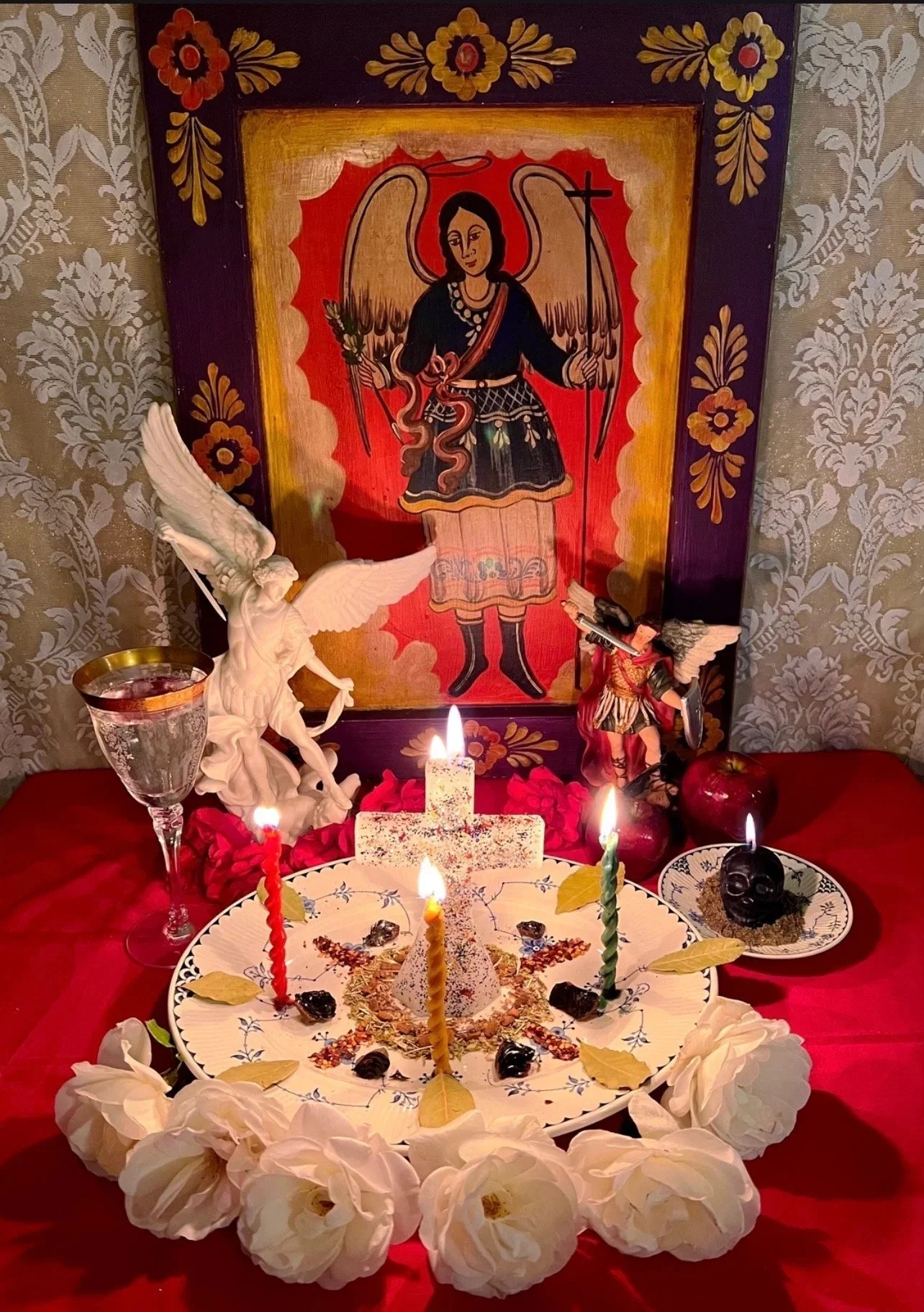 A Día de los Muertos altar with candles, skulls, flowers, and traditional decorations, including a framed image of an angel or saint in the background, surrounded by decorative items such as figurines and a chalice.