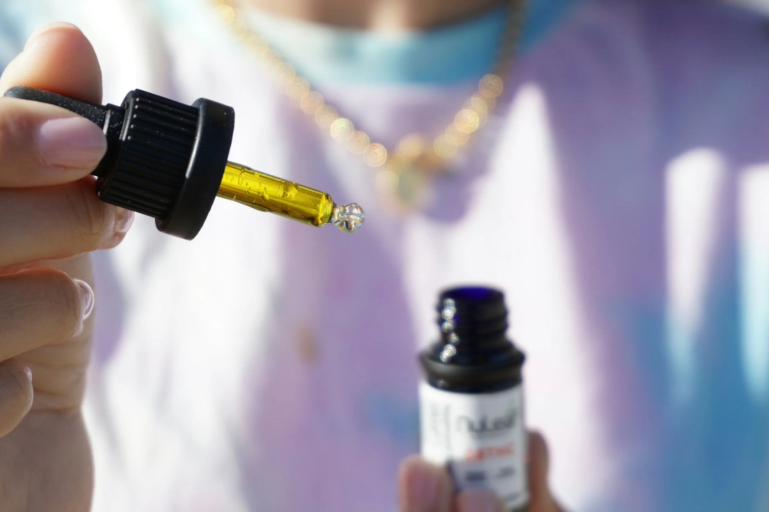 Close-up of a hand holding a small bottle of liquid medication with a dropper, with a blurred background showing another small bottle.