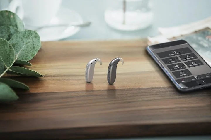 A smartphone on a wooden desk displays a remote control app, with two hearing aids placed nearby, and a green plant on the left side.