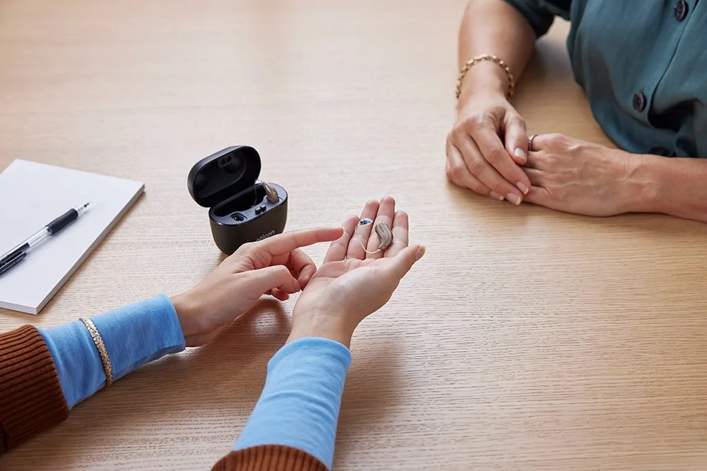 Person handing over hearing aids to another person at a wooden table with a notebook, pen, and hearing aid case.