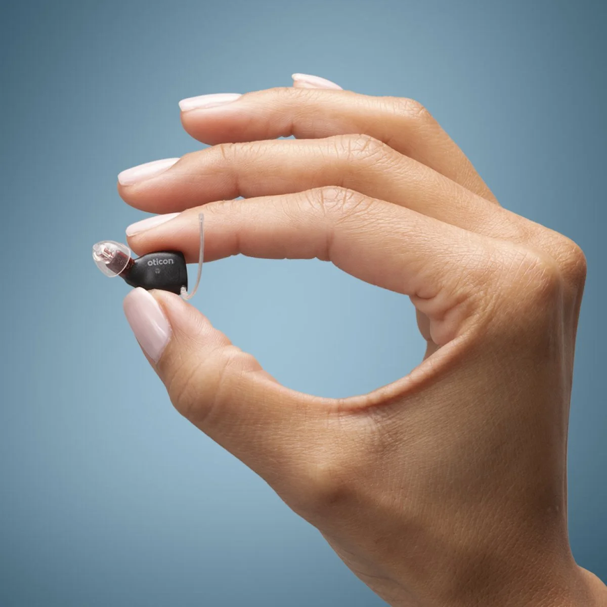 A hand holding a small hearing aid between the thumb and index finger against a blue background.