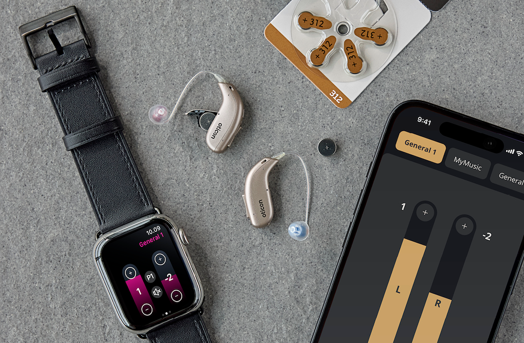 Smartwatch displaying a music control app, hearing aids, a pack of hearing aid batteries, and an iPhone with a music volume adjustment screen on a gray surface.