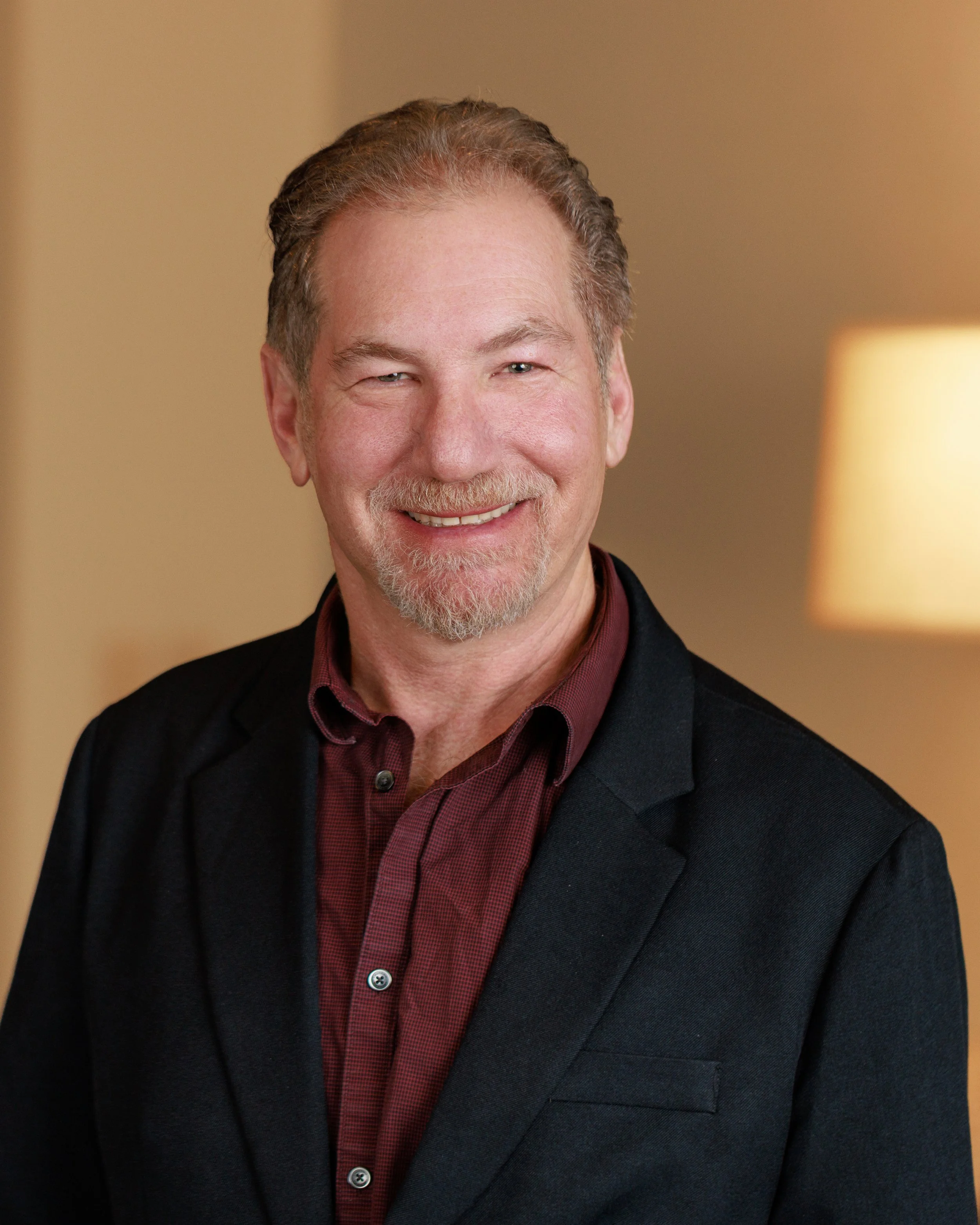 Portrait of a middle-aged man with gray hair, a mustache, and a goatee, smiling and wearing a dark blazer over a maroon shirt, in a warmly lit room.