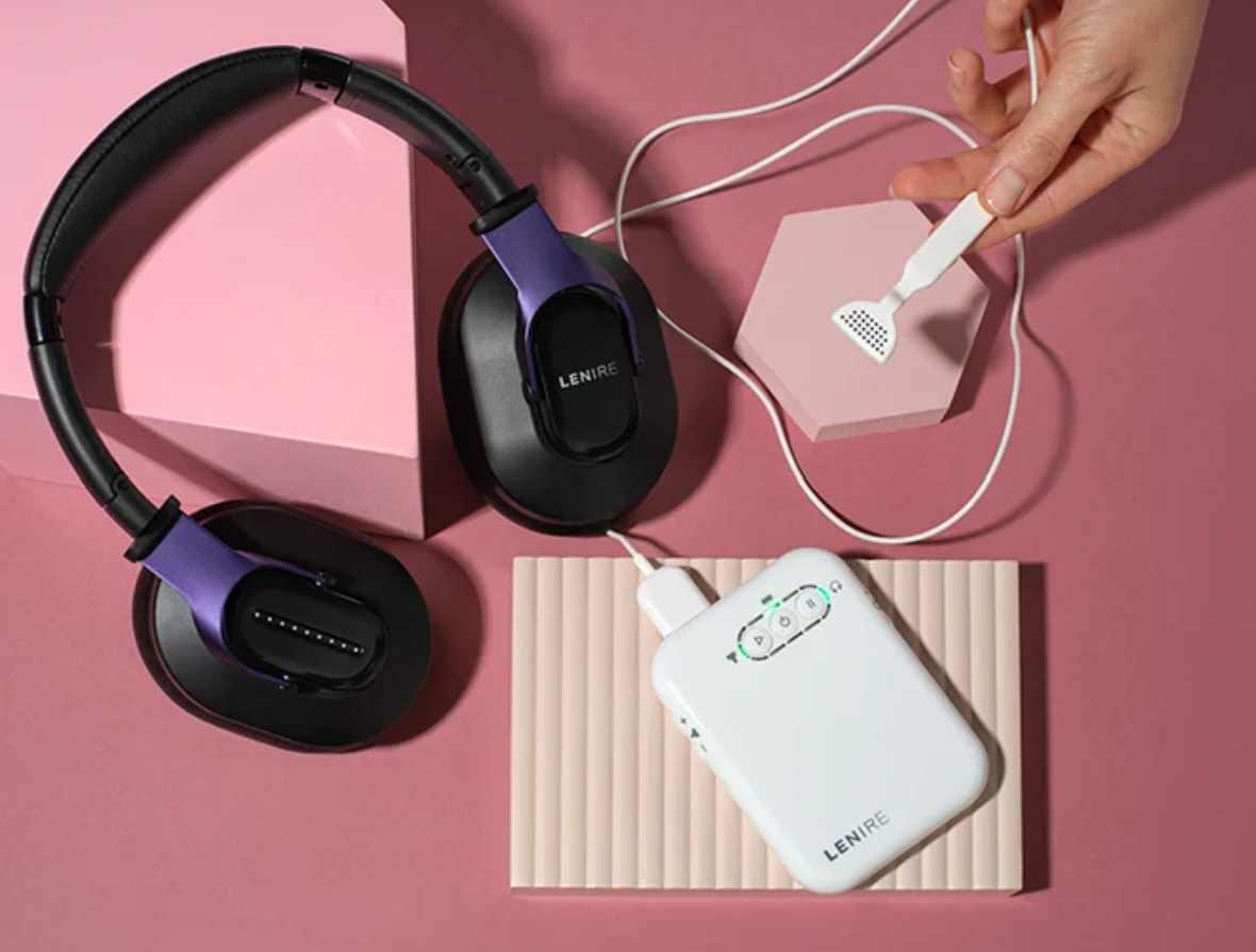 A person holding a white cleaning brush near a pink hexagonal object, with black and purple wireless headphones, a white portable device, and a pink box arranged on a pink surface.