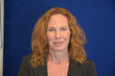 A woman with curly red hair wearing a black blazer stands against a blue background.