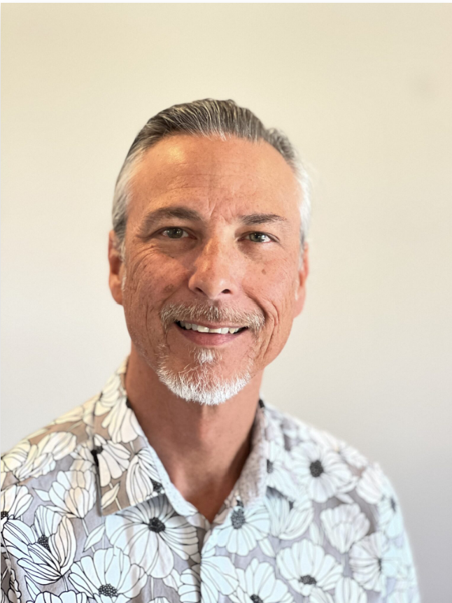 A middle-aged man with gray hair and a goatee smiling, wearing a floral-patterned shirt, standing against a plain light-colored wall.