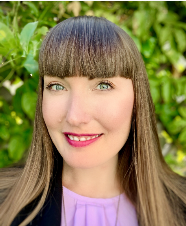 Close-up portrait of a woman with long brown hair, bangs, green eyes, and pink lipstick, smiling outdoors with green leafy background.