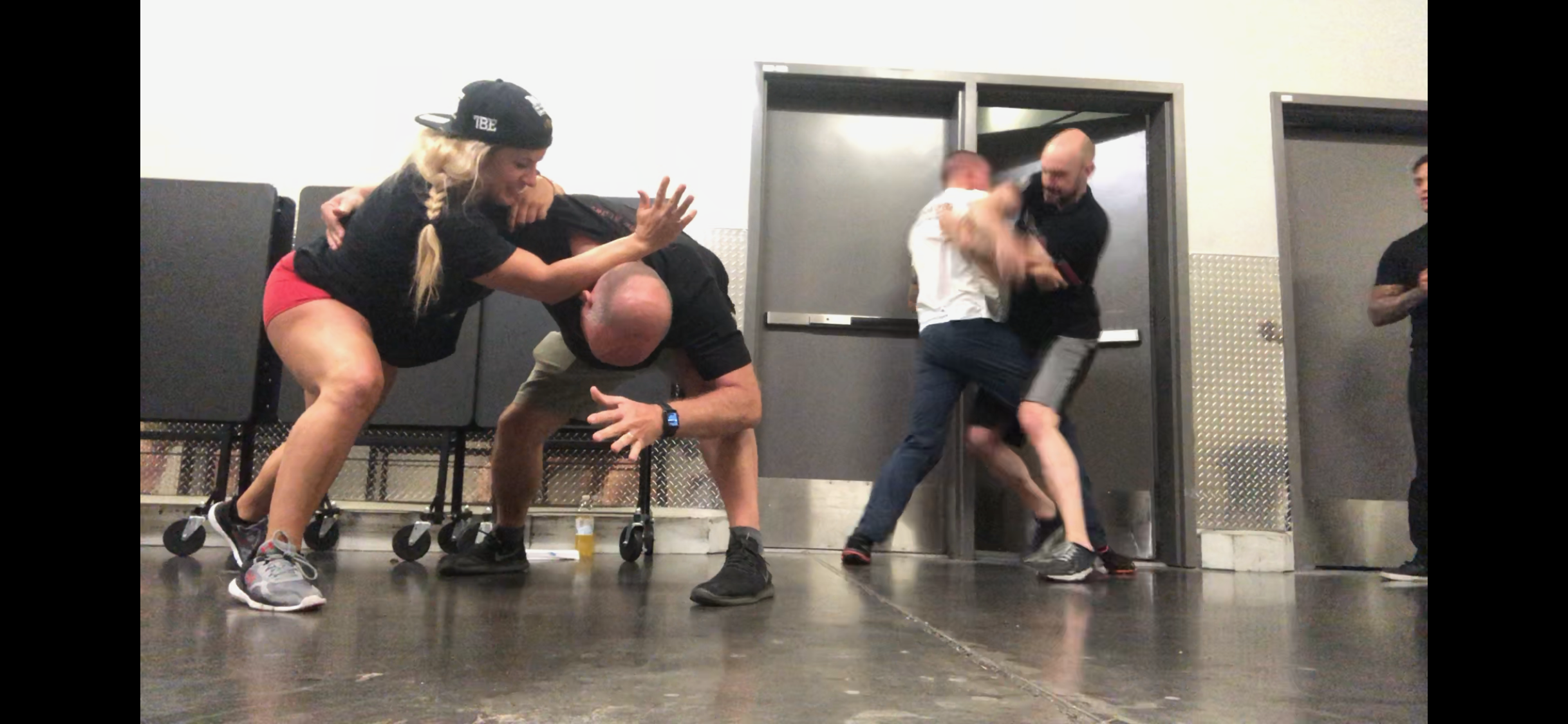 Women's empowerment expert Dr. Alice Atalanta engaging in a physical self-defense practice with a group of men, with two separate pairs grappling in a room with metal doors.