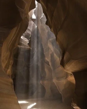 Sunlight streaming into a narrow sandstone slot canyon, creating dramatic light beams and shadows on the rock walls.