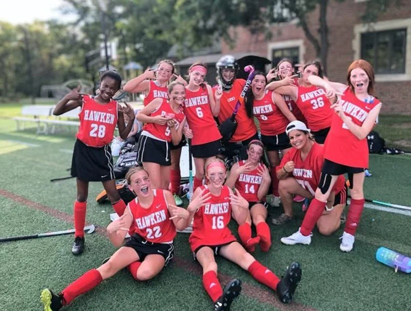 A group of female field hockey players posing enthusiastically on a sports field with their field hockey coach, women's empowerment expert Dr. Alice Atalanta.