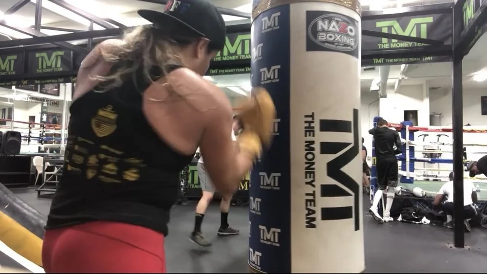 Women's empowerment expert Dr. Alice Atalanta practicing boxing with a punching bag in a gym, surrounded by "The Money Team" branding, feeling like an empowered woman in a male dominated environment.