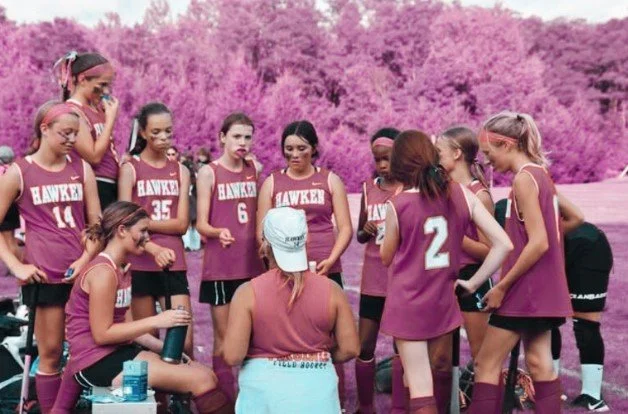 Female sports team in maroon jerseys with their coach, women's empowerment expert Dr. Alice Atalanta, having a discussion on a field.