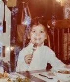 Women's empowerment expert Dr. Alice Atalanta as a young child smiling and holding a fork at a dining table, wearing a white shirt, a girl feeling joyful, free, and comfortable in her own skin.