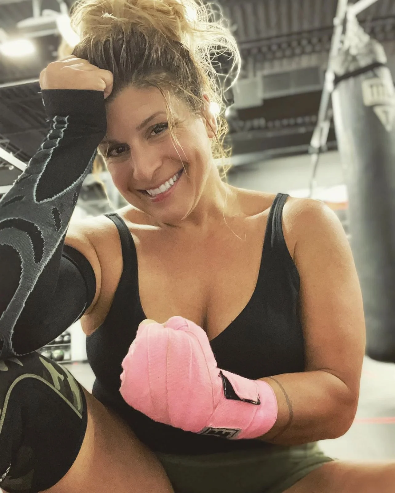 Smiling women's empowerment expert Dr. Alice Atalanta,  in workout attire and pink hand wraps at a gym where she has been practicing combatives for self-defense, sitting in front of a punching bag feeling empowered and strong.