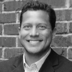 Man, a US Army Green Beret named Brent Cooper, smiling in front of brick wall, wearing a suit jacket and checkered shirt while he served as Executive Director of the Green Beret Foundation.