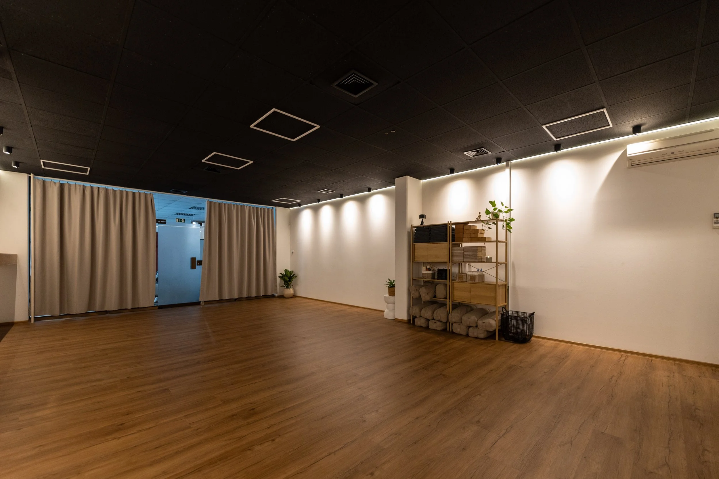 Interior of a modern room with hardwood floors, black ceiling with square recessed lighting, white walls, beige curtains, potted plants, and a wooden shelf with boxes and rolled-up mats.