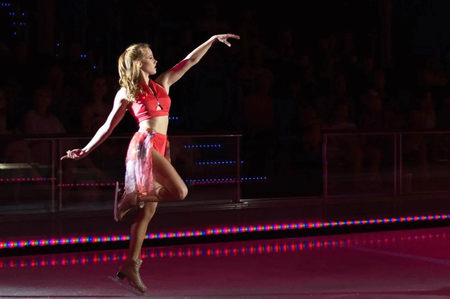 A female professional figure skater dressed in an orange top and sheer skirt is on the ice, balancing on one leg with arms outstretched in a skating routine. While she is lit up by the spotlight, the background is dark with an audience watching.