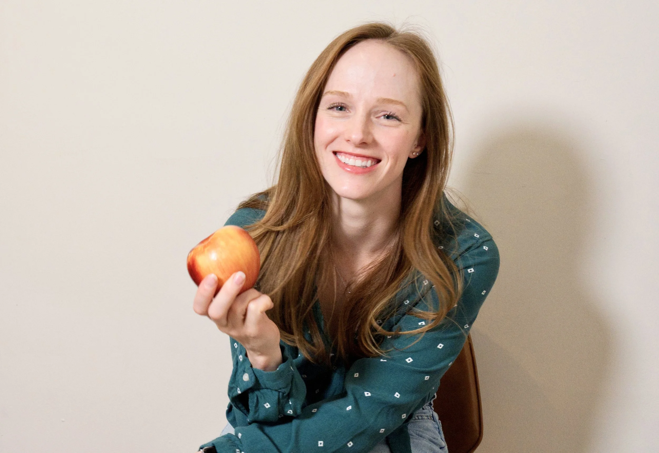 A woman with long red hair is smiling and holding a red apple in her hand, seated against a plain beige wall.