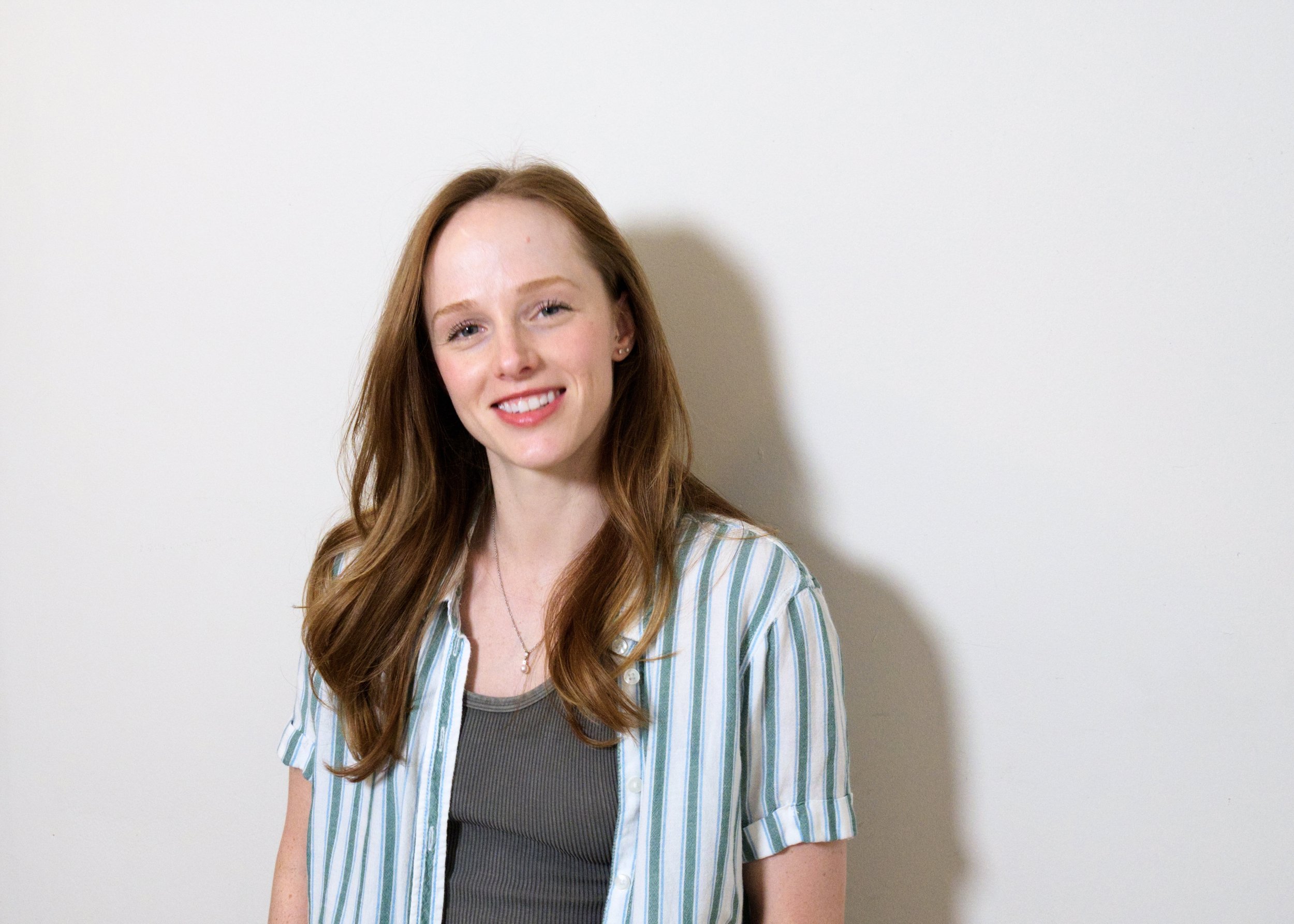 Young woman with long red hair smiling, wearing a striped shirt over a green top, standing against a plain light-colored wall.