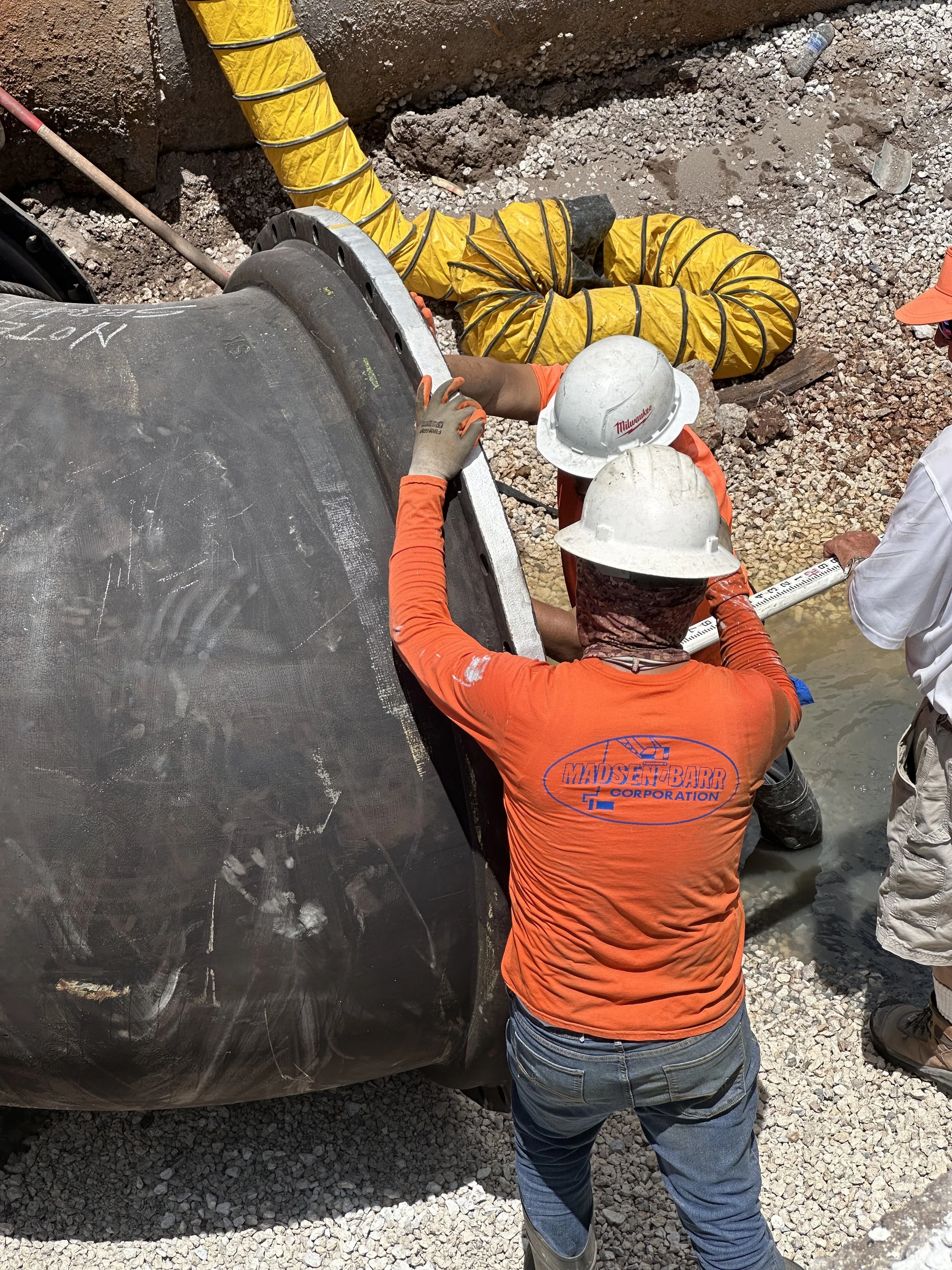 Construction workers installing or inspecting a large underground pipe at a construction site. One worker is wearing an orange shirt with a logo and a white hard hat, while the other has a white hard hat and is holding a measuring tape.