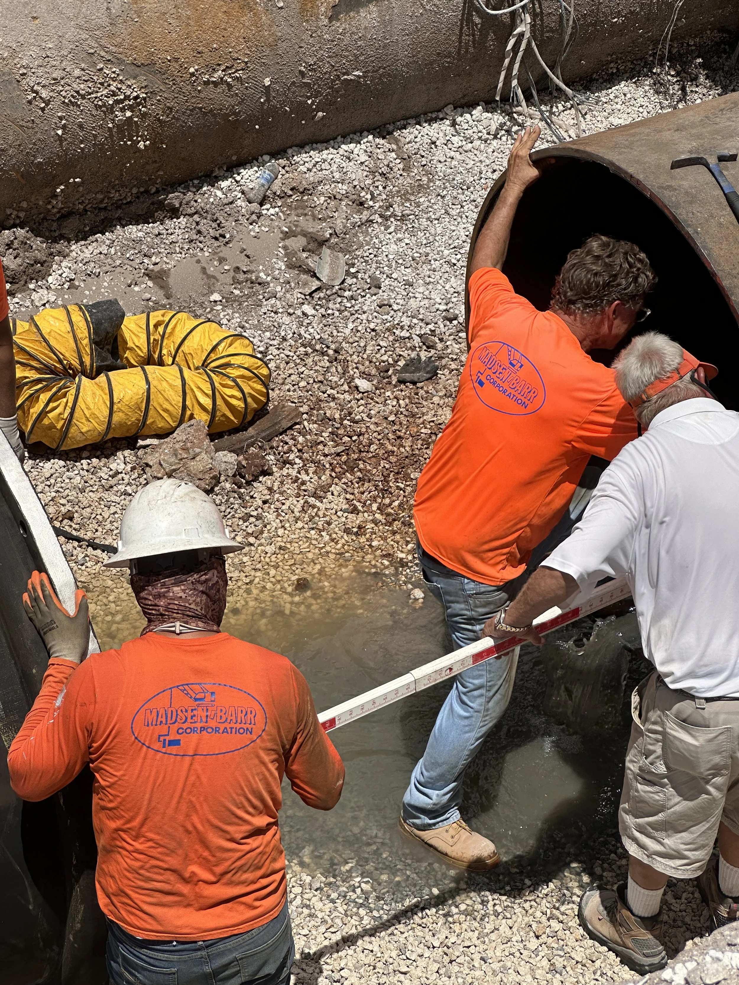 Workers inspect a small water stream using a measuring tape, with construction tools and safety gear nearby.