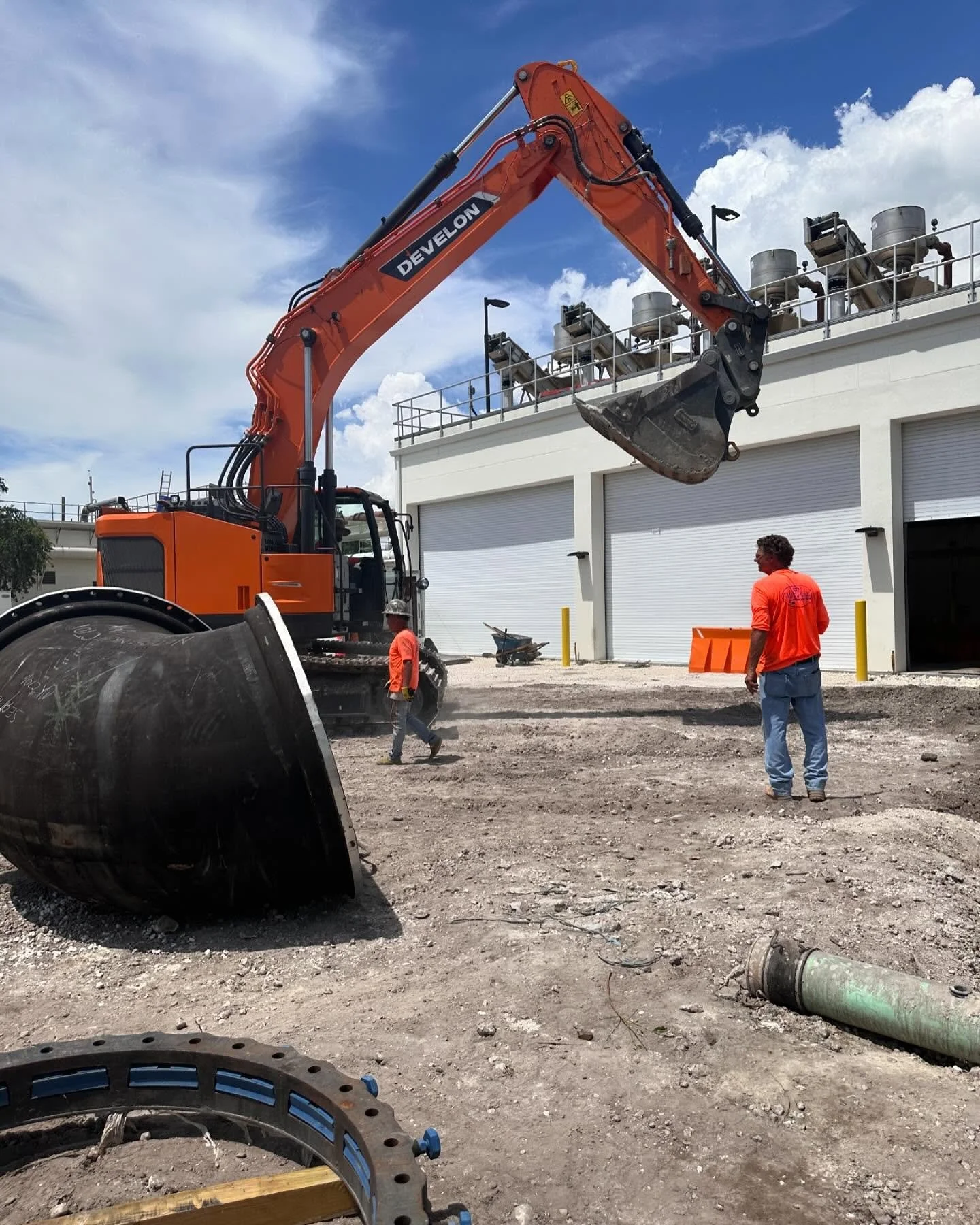 Construction site with an orange excavator lifting materials, two workers in orange shirts walking, a large black pipe on the ground, and a building with a white garage door in the background.