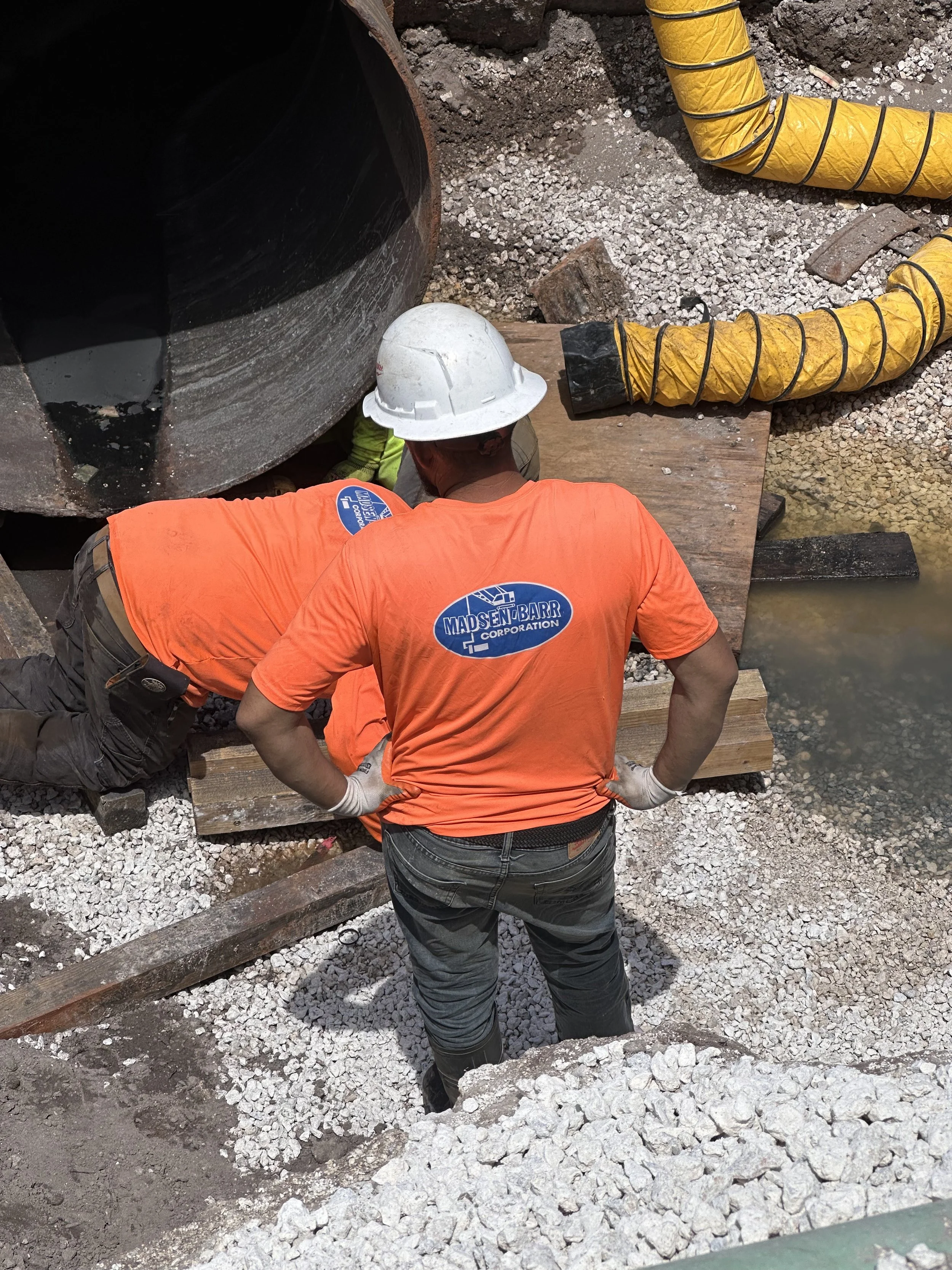 Two construction workers wearing orange shirts and safety helmets working on a large pipeline underground. One worker is kneeling and the other is standing, both are surrounded by gravel and wooden planks. Yellow flexible hoses are nearby.