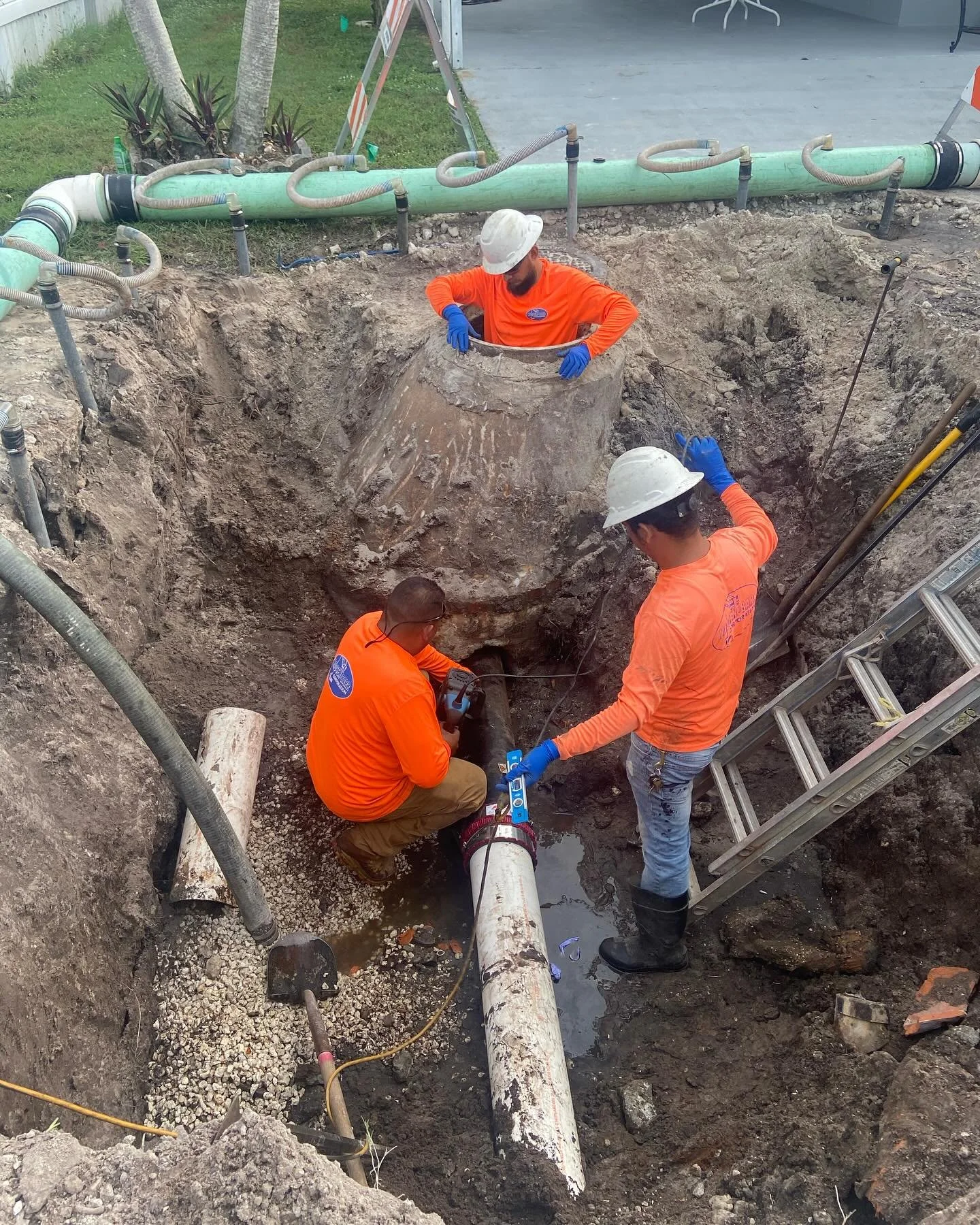 Three workers in orange shirts and white hard hats repairing underground pipes in a construction site. One worker is holding a pipe, another is working on the pipe with tools, and the third is observing. The trench exposes large pipes and construction equipment around.
