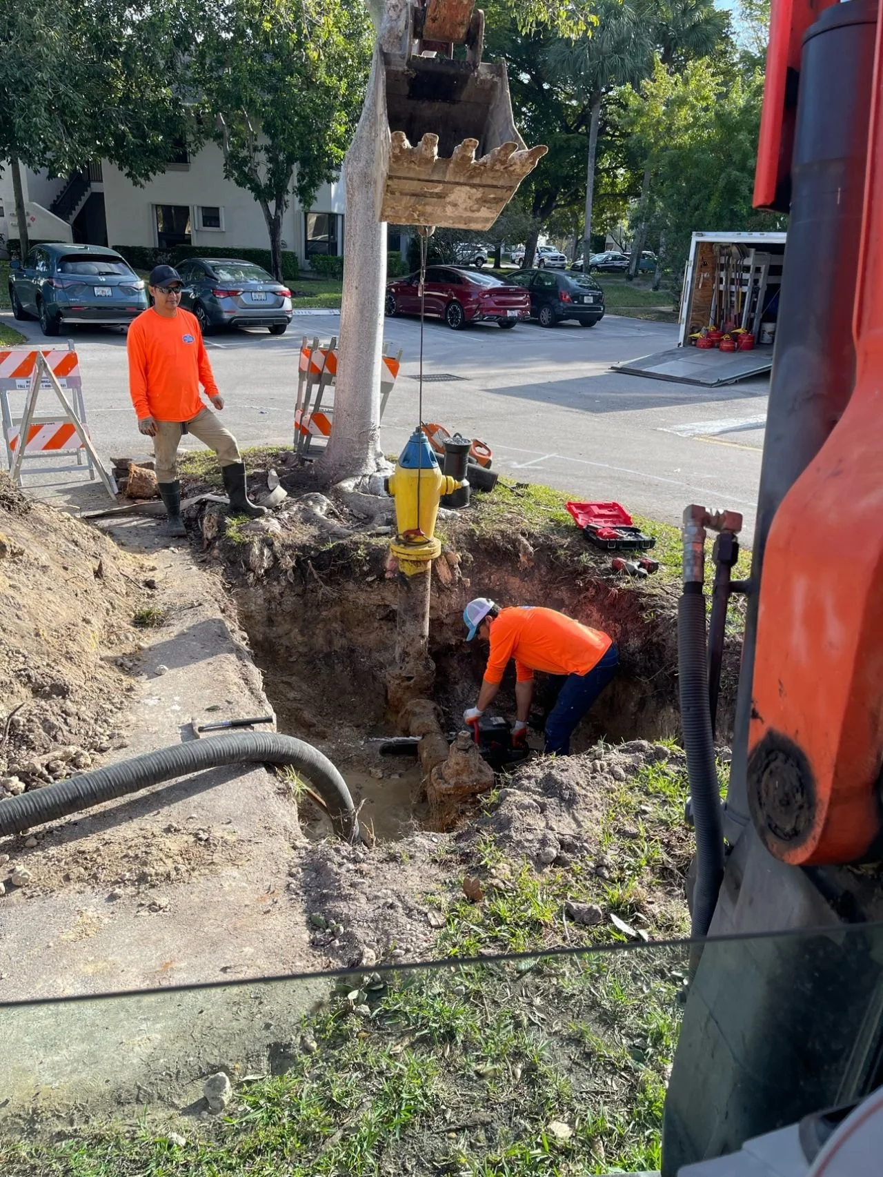 Workers in orange shirts repairing underground water pipes in a trench near a parking lot, with equipment and safety barriers nearby.