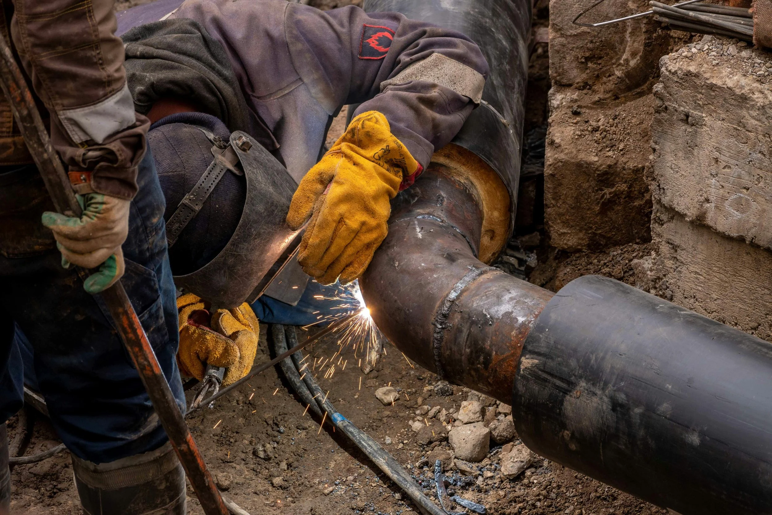 Worker welding large black pipe in an excavation site, sparks flying, wearing yellow gloves and a dark protective jacket.