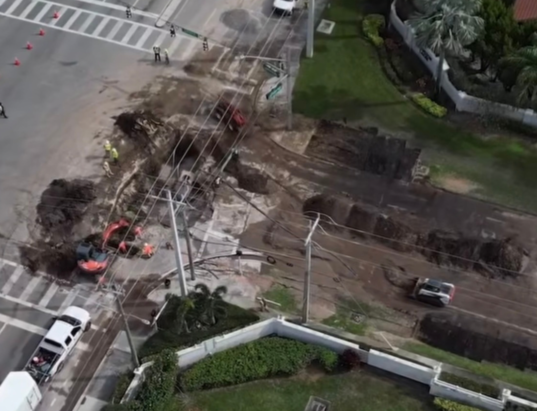 A large section of road is damaged with a collapsed utility pole and exposed wires. Construction workers in yellow vests are on site, and multiple cars are present nearby, some involved in the accident. Traffic cones are placed around the area.