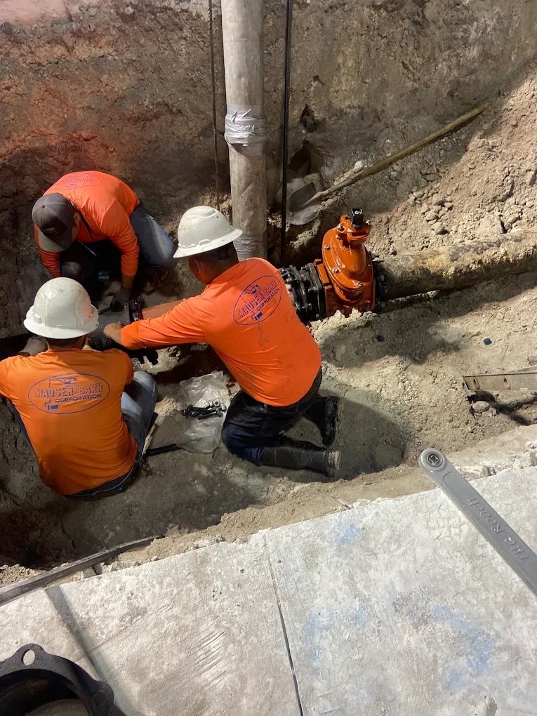 Three construction workers in orange shirts and white helmets working on a plumbing connection in a trench.