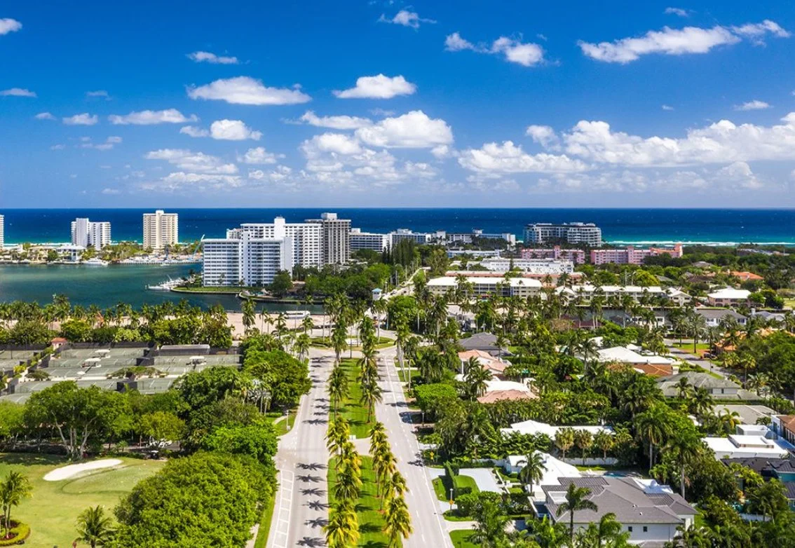 Aerial view of a coastal city with high-rise buildings, a river, houses, palm trees, and the ocean under a partly cloudy sky.