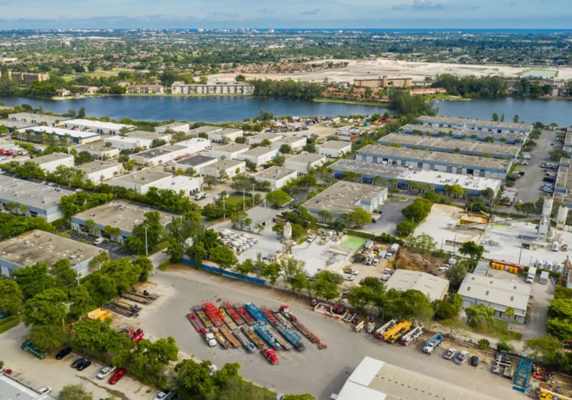 Aerial view of an industrial area near a river, with warehouses, parking lots, and storage yards with trucks and construction equipment, and a cityscape in the background.