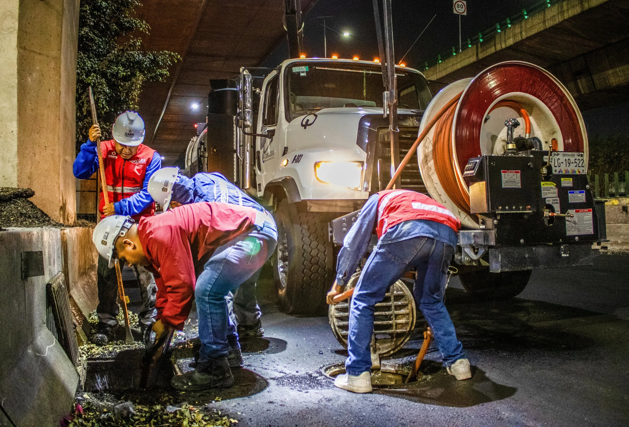 Workers in safety helmets and vests repairing a road under a bridge at night, with construction equipment and a large spool of cable nearby.